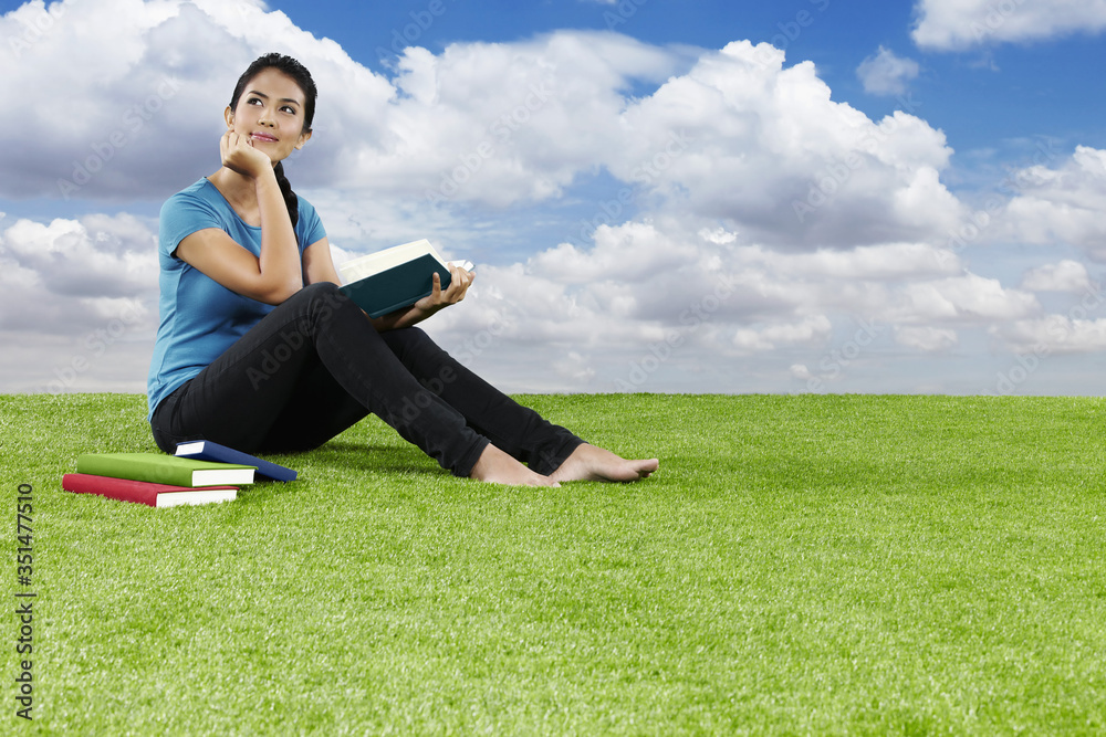 Fototapeta premium Young woman sitting on grass reading a book