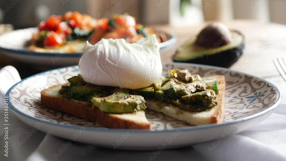Close-up view cutting perfect poached eggs with avocado paste on crispy toast, fresh green avocado spread sandwiches recipe in restaurant as healthy breakfast.
