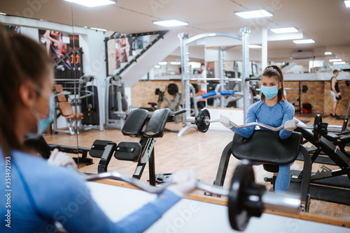A beautiful caucasian girl with a mask and gloves stands and exercises with weights in the gym. COVID - 19 virus protection