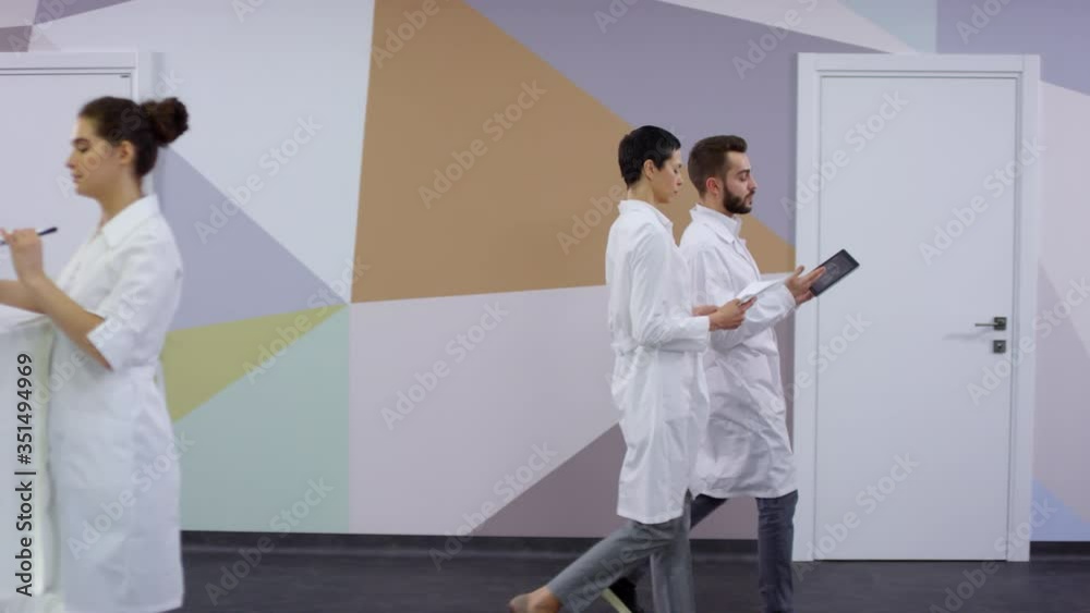 PAN of male doctor in white coat holding tablet and discussing work with female colleague while walking along hallway in hospital. Young female medical specialist carrying clipboard to reception area PAN of male doctor in white coat holding tablet and discussing work with female colleague while walking along hallway in hospital. Young female medical specialist carrying clipboard to reception area