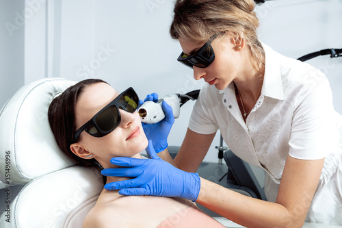 Woman Receiving Facial Beauty Treatment Removing Pigmentation At Cosmetic Clinic. Intense Pulsed Light Therapy Rejuvenation. Laser cosmetology blond doctor and patient wearing black protection glasses