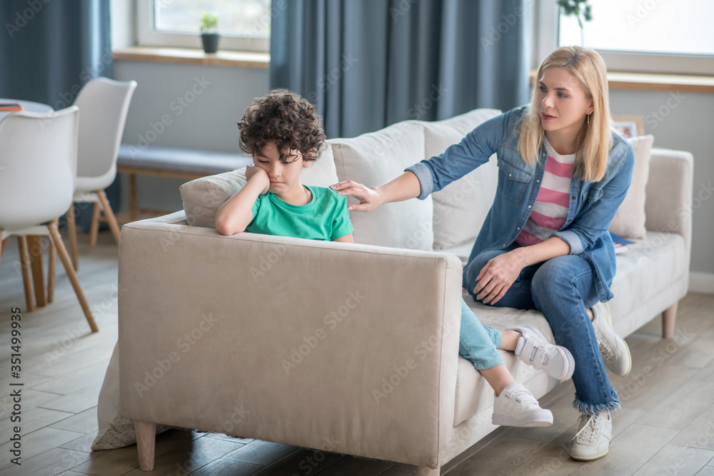Sad curly boy sitting on sofa, blonde female touching his shouder, trying to cheer him up