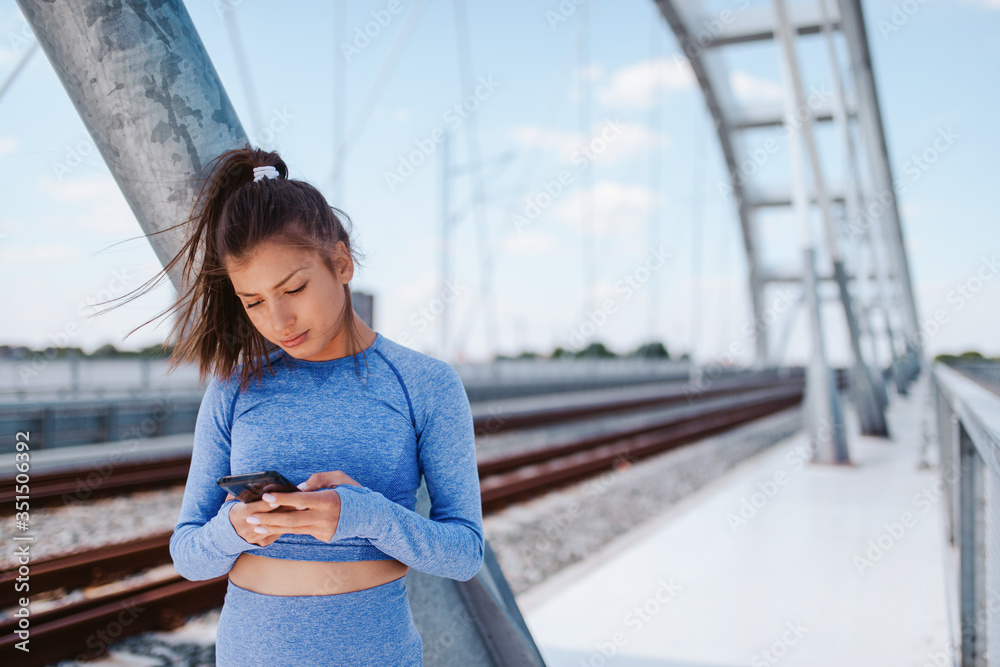 Portrait of a young beautiful caucasian girl in blue sports equipment with a phone in her hand on the bridge