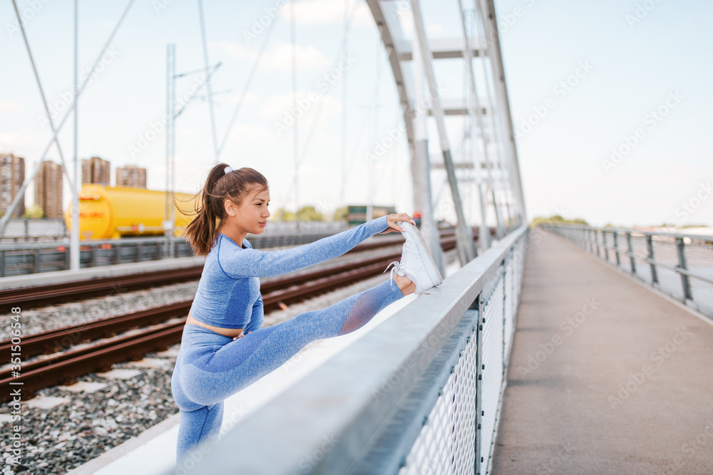 A young beautiful caucasian girl in blue sports equipment stretches her leg on the bridge. Training and healthy life