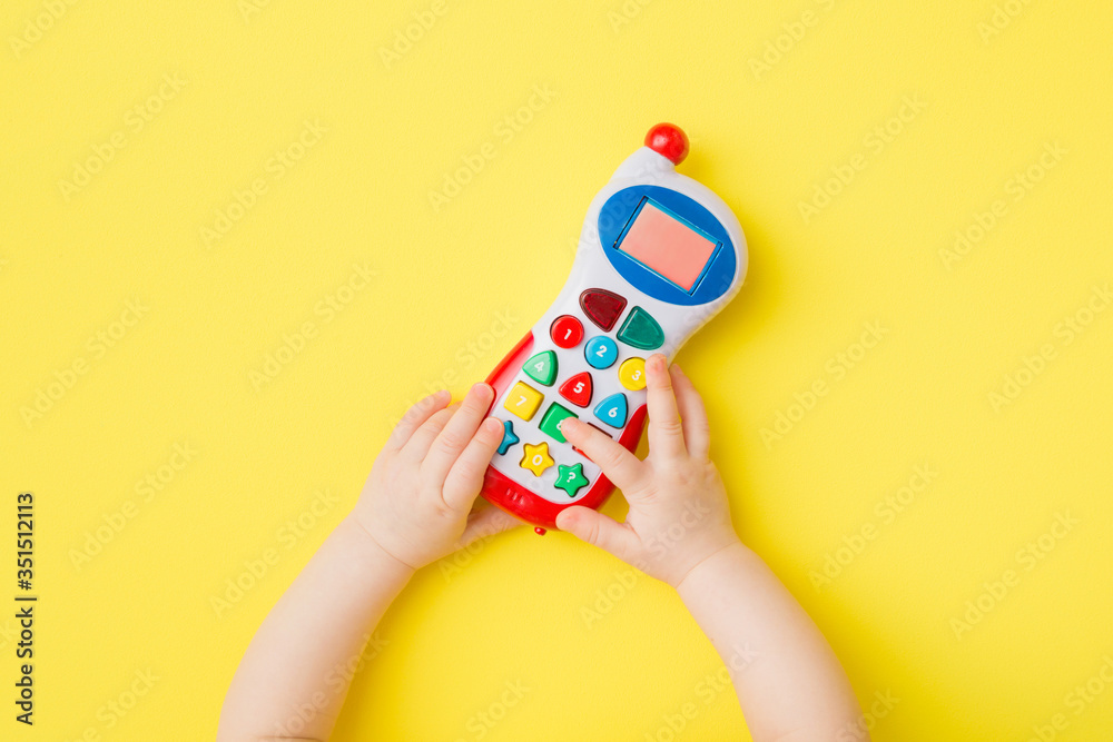 Obraz premium Baby hands playing with colorful toy of mobile phone on bright yellow table background. Closeup. Point of view shot. Top down view.