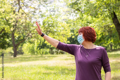 Aged, matured woman in medical mask welcomes friends from afar, keeping social distance.