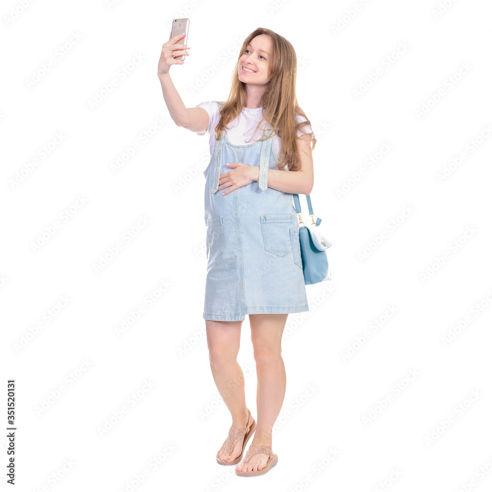 Pregnant woman in denim sundress with blue bag standing looking smiling, making selfie on smart phone on white background isolation Pregnant woman in denim sundress with blue bag standing looking smiling, making selfie on smart phone on white background isolation