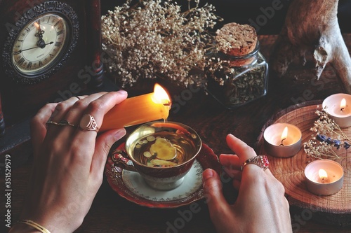 Wiccan witch wearing vintage jewelry holding yellow candle and pouring wax into a red gold vintage teacup as a divination. Reading Candle Wax - Carromancy, Ceroscopy among nature items dried flowers photo