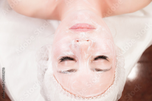A young attractive girl in a cosmetologist s office with her face covered in a cosmetic rejuvenating white mask with a brush applied lies and relaxes. Facial skin care concept