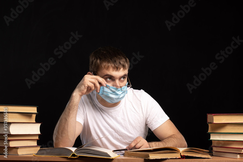 young man in a medical mask at a table with books on a background of black wall. looks away with his glasses down. student is in quarantine. distance learning. office employee