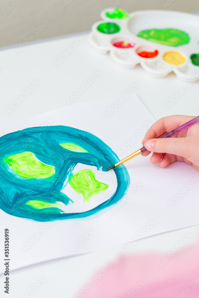 Close up of child's hand drawing planet Earth with a brush by acrylic paints on white paper. Earth day celebration.