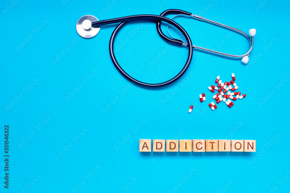 word addiction in woods next to lozenges and stethoscope on a blue background word addiction in woods next to lozenges and stethoscope on a blue background