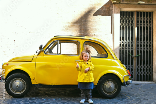 little cute girl 3 years old in a yellow raincoat against a yellow car in Rome Italy Via Margutta smiling shows class photo