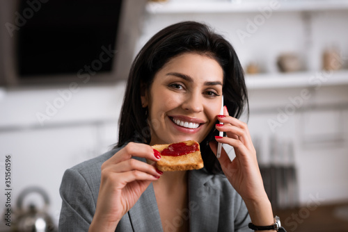 cheerful businesswoman holding toast bread with jam while talking on smartphone and hiding problem of domestic violence