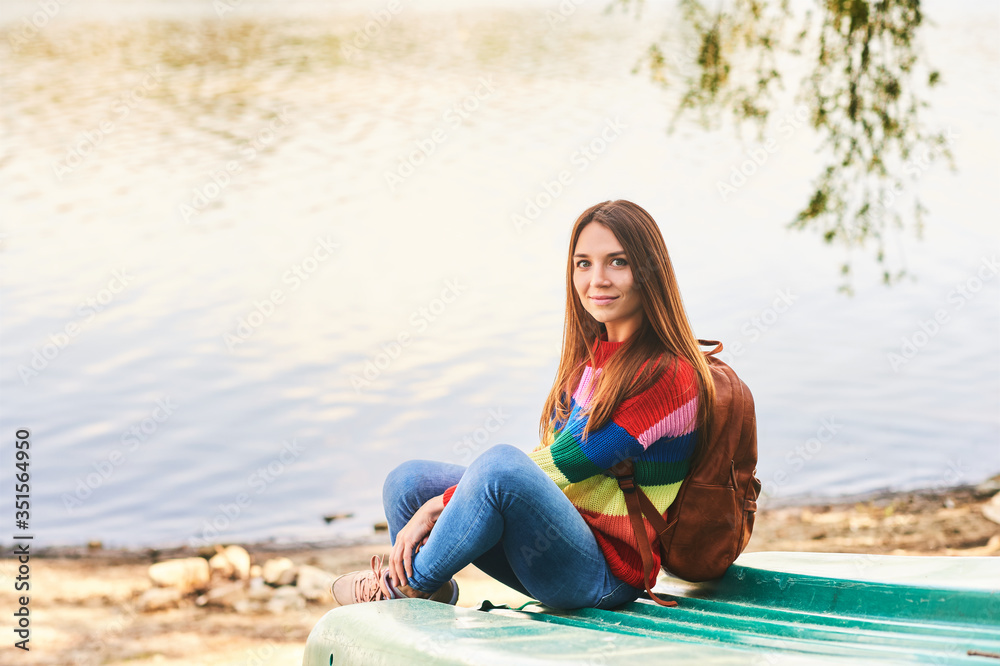 Outdoor portrait of young beautiful woman relaxing by river on a nice warm day, wearing colorful pullover and backpack