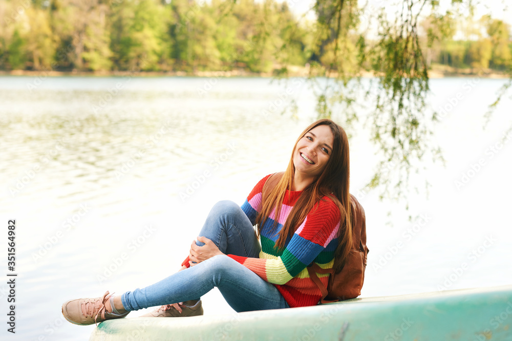 Outdoor portrait of young beautiful woman relaxing by river on a nice warm day, wearing colorful pullover and backpack