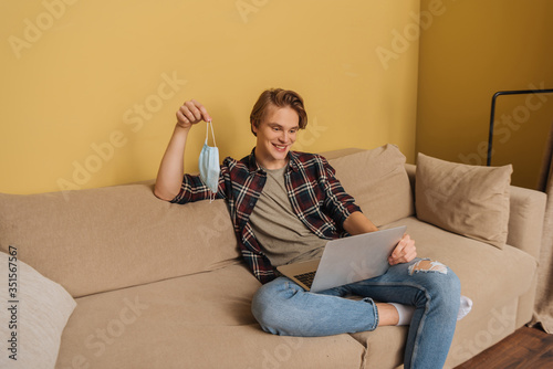 cheerful freelancer holding medical mask near laptop in living room, end of quarantine concept