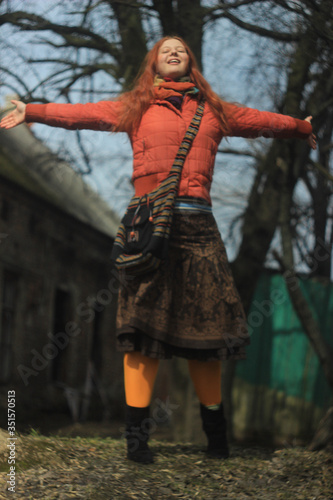 A happy red-haired young girl in a bright red jacket and pantyhose  in a skirt with floral patterns  poses against the backdrop of nature