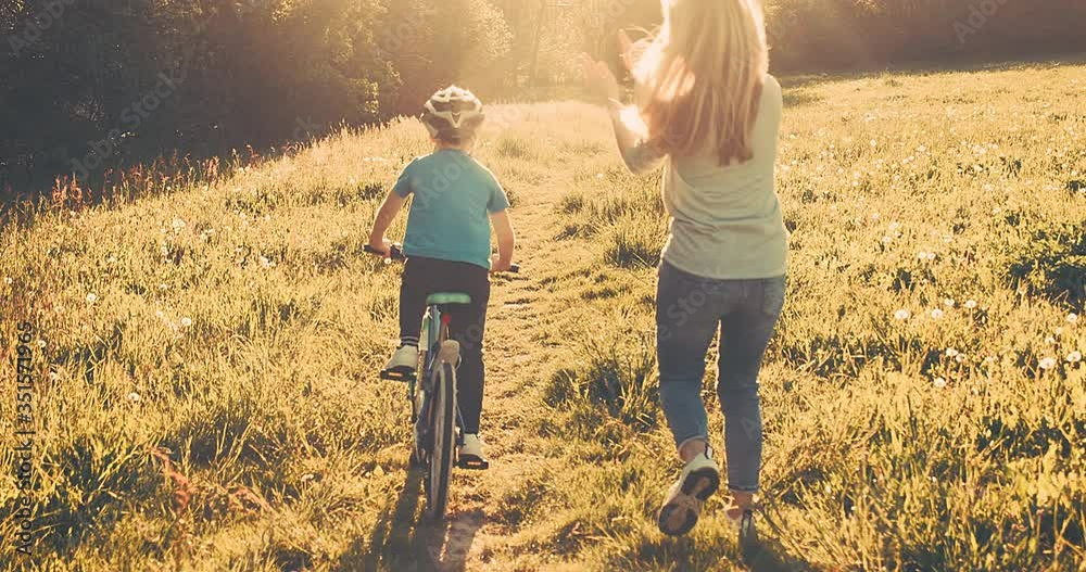 Mother teaching son to ride bicycle. Happy cute boy in helmet learn to riding a bike in park on green meadow in summer day at sunset time. Family weekend. 4K video Slow motion