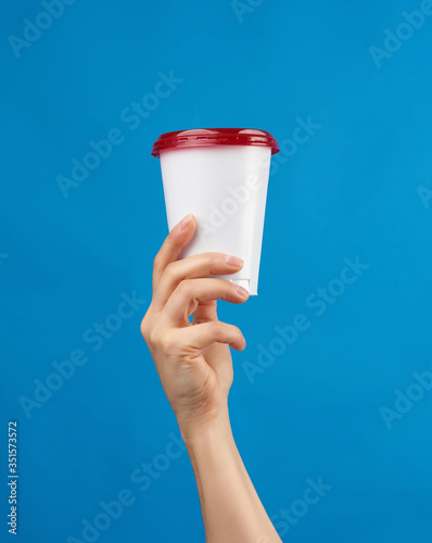 female hand holds a disposable white glass with a red cap on a blue background