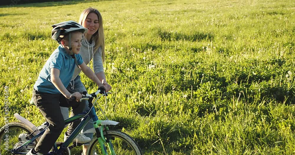 Mother teaching son to ride bicycle. Happy cute boy in helmet learn to riding a bike in park on green meadow in summer day at sunset time. Family weekend. 4K video Slow motion