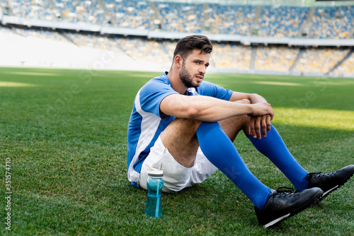 professional soccer player in blue and white uniform sitting on football pitch with sports bottle at stadium