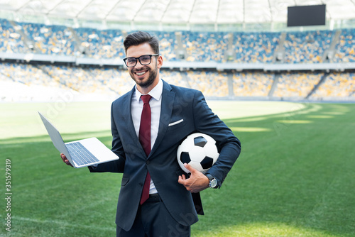 Fototapeta smiling young businessman in suit with laptop and soccer ball sitting on footbal
