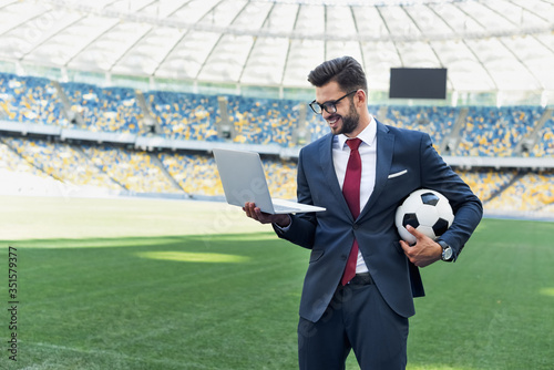 Fototapete smiling young businessman in suit with laptop and soccer ball at stadium, sports