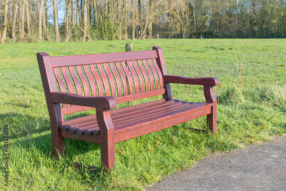 Brown wooden bench on the side of an abandoned path with a green meadow in the background Brown wooden bench on the side of an abandoned path with a green meadow in the background