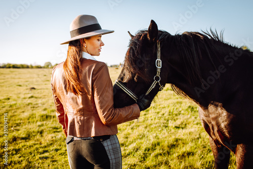 Beautiful smiling young woman in a hat and gloves playing with a broun horse in a field on a sunset. Horseback riding.