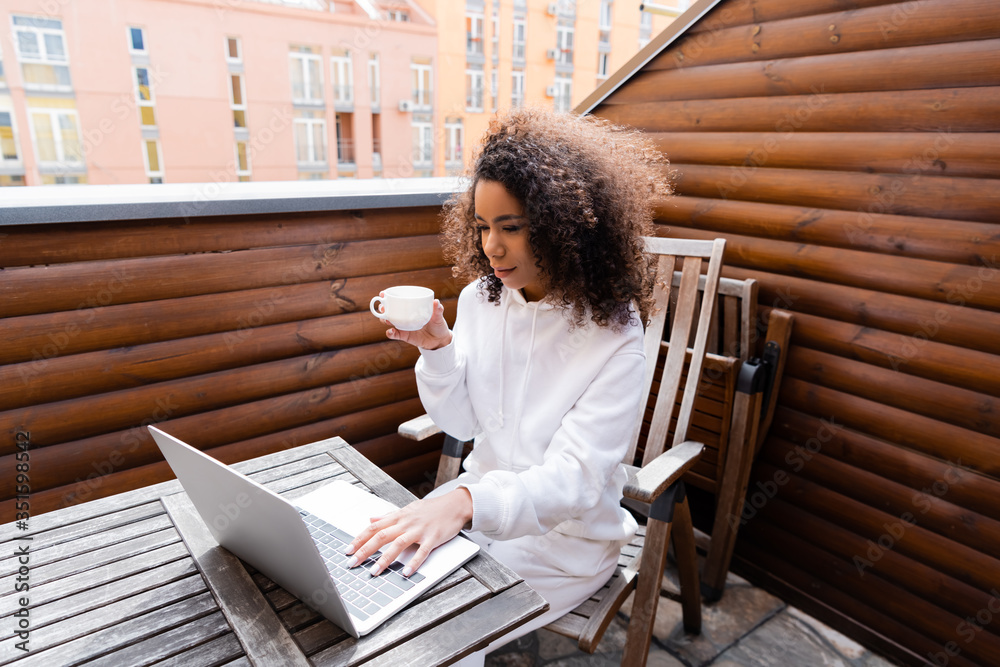 curly african american freelancer holding cup and working outside - obrazy, fototapety, plakaty 