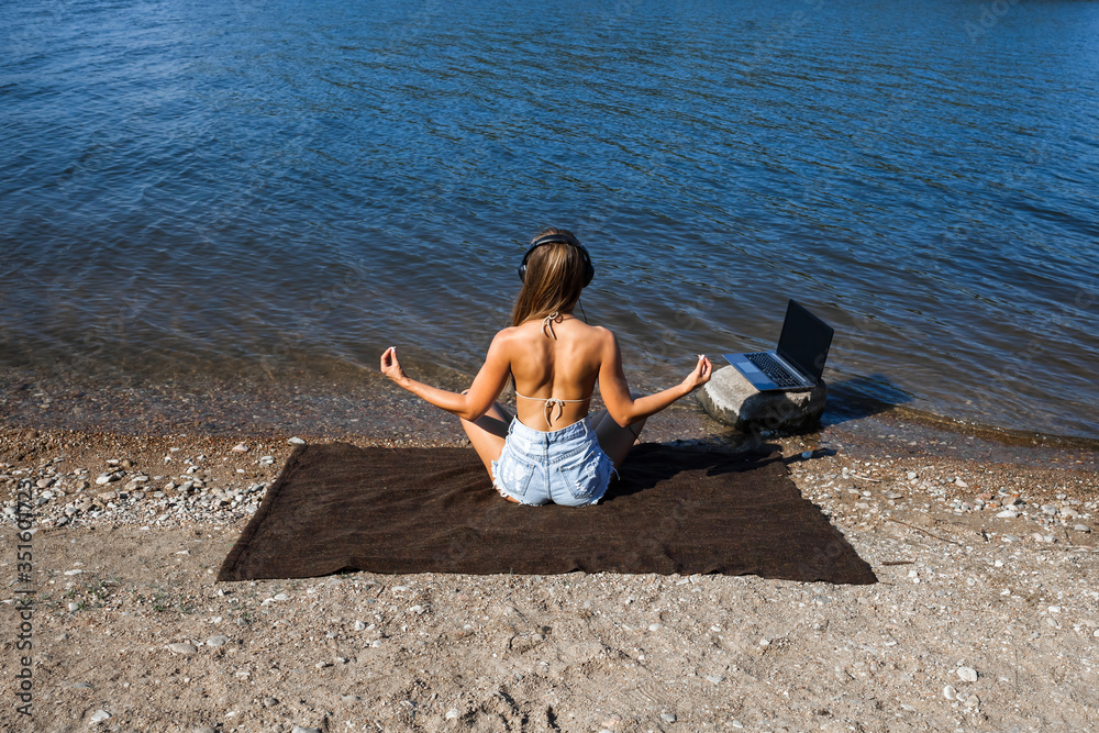 A young woman with blond hair is relaxing and resting on the sandy shore, listening to music on the lake. The girl practices yoga. A young woman with blond hair is relaxing and resting on the sandy shore, listening to music on the lake. The girl practices yoga.