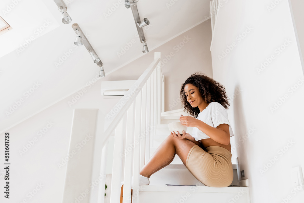 happy african american woman sitting on stairs and holding bowl and spoon happy african american woman sitting on stairs and holding bowl and spoon
