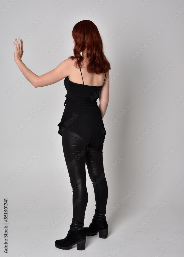 portrait of a pretty girl with red hair wearing black leather pants and top. Full length standing pose isolated against a grey studio background portrait of a pretty girl with red hair wearing black leather pants and top. Full length standing pose isolated against a grey studio background
