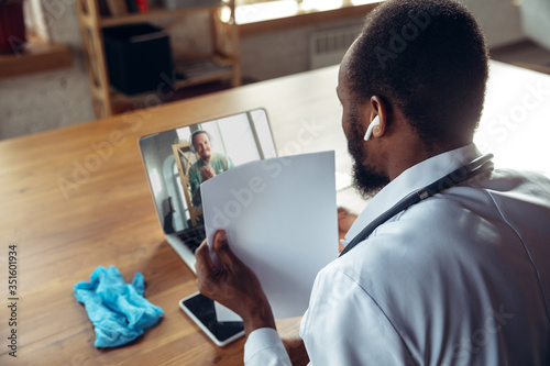 Doctor advising the patient online with laptop. African-american doctor during his work with patients, explaining recipes for drug. Daily hard work for health and lives saving during epidemic. photo