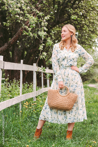 Young beautiful woman retro model posing with straw handbag and smile in summer park. Happy attractive hippie girl in white retro floral dress walk on green countryside garden. Enjoy nature outdoors