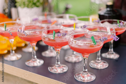 Pink alcoholic cocktail in sour glass with sprig of lavender pinned at it's rim with decorative clothespin standing in row at outdoor cocktail party
