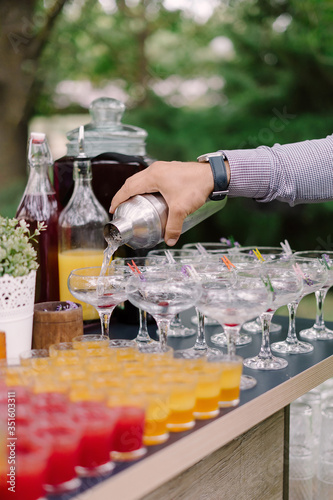 Bartender pouring cocktails from stainless steel cocktail shaker into sour glasses, near by rows of orange and red coloured shooter drinks at open air cocktail party