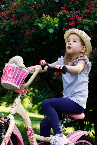 Little happy girl sitting on a bicycle and playing. She is wearing a hat and a T-shirt. Summer warm day. Flowering bush on a background