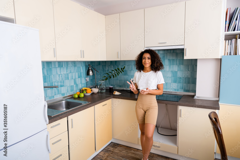 cheerful african american girl holding cup and showing peace sign near fresh fruits