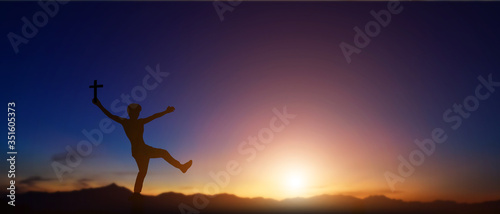 Silhouette of man holding christian cross in hands at sunset background.