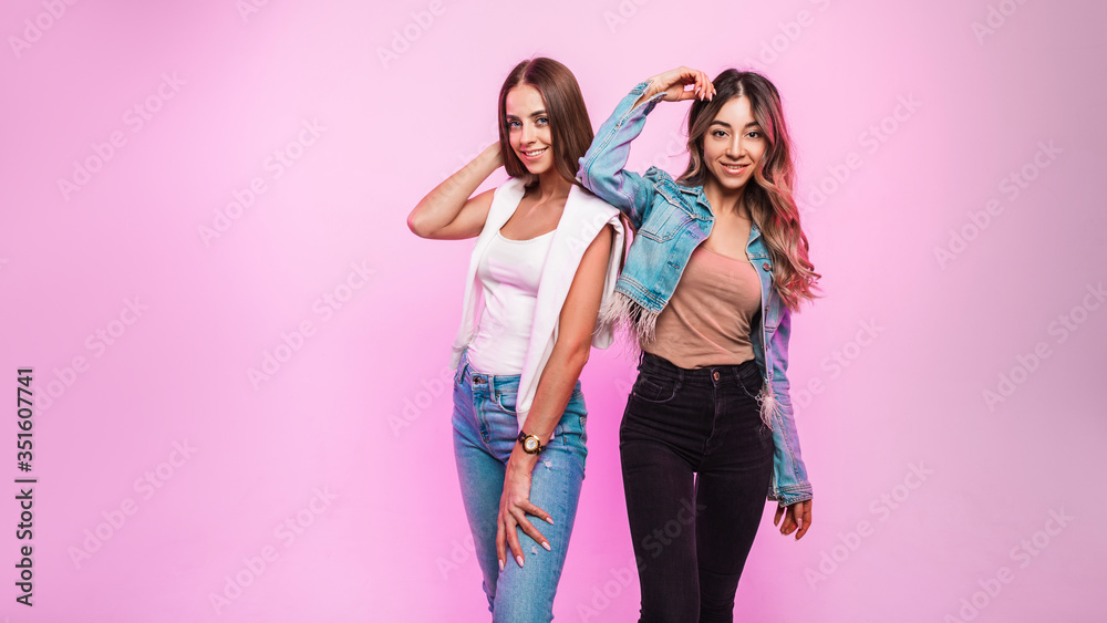 Two cute happy women with charming smiles in trendy youth jeans clothes posing and smiling on  background of pink wall in the room. Pair attractive positive girlfriends in denim wear in the studio. - obrazy, fototapety, plakaty 