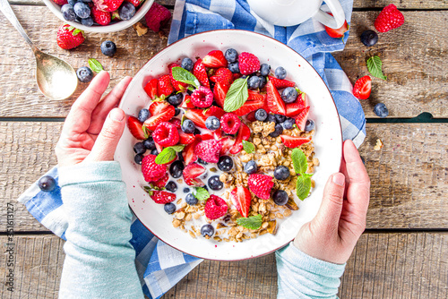 Healthy morning food, Breakfast oatmeal granola or muesli with various berry and milk, yogurt, wooden rustic background. Woman hands hold plate, sppon, flatlay top view photo