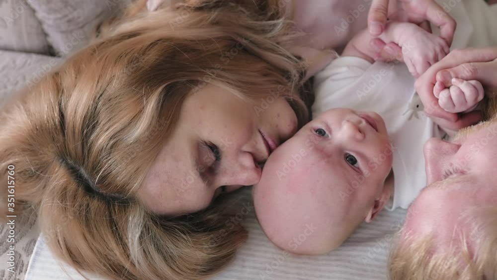 Happy young family with a newborn baby. Mom, dad and newborn lying on the bed, top view. Happy mom and dad kiss and hug the baby. Parenthood concept. Happy young family with a newborn baby. Mom, dad and newborn lying on the bed, top view. Happy mom and dad kiss and hug the baby. Parenthood concept.