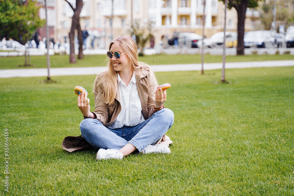 Beautiful, attractive, young caucasian girl with long blond hair sitting on the grass in a spring park. Woman holding donuts in her hands. Smiling and having a good time in nature outdoors. Posing Beautiful, attractive, young caucasian girl with long blond hair sitting on the grass in a spring park. Woman holding donuts in her hands. Smiling and having a good time in nature outdoors. Posing