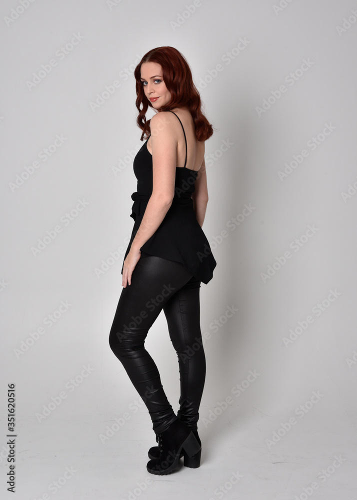 portrait of a pretty girl with red hair wearing black leather pants and top. Full length standing pose in side profile isolated against a grey studio background portrait of a pretty girl with red hair wearing black leather pants and top. Full length standing pose in side profile isolated against a grey studio background