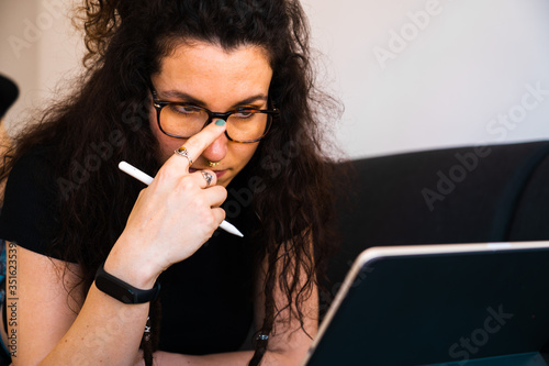 Young brunette girl working from home with her tablet with a digital pencil while touching her eyeglasses lying on a black couch