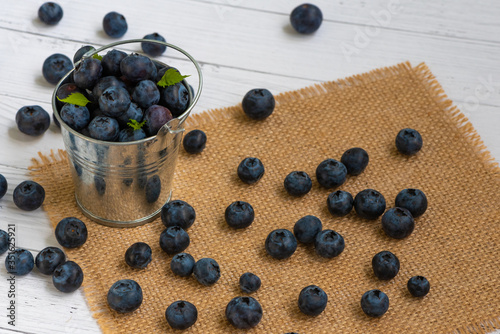 Close-up of blueberries in a small bucket with green leaves and scattered on a jute napkin on a light table.