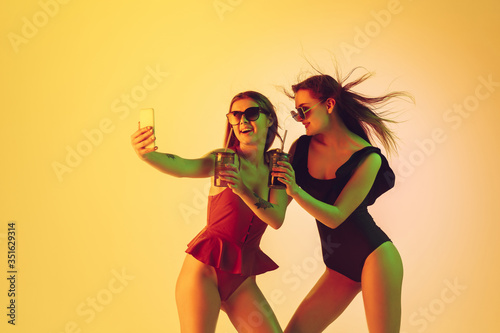 Beautiful young girls' portrait isolated on yellow studio background in neon light. Women in fashionable bodysuits. Facial expression, summer, weekend, beauty, resort concept. Vacations, youth.