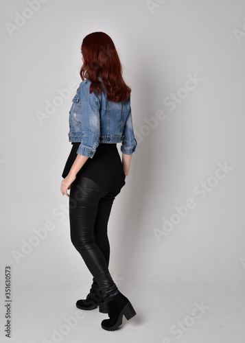 portrait of a pretty girl with red hair wearing black leather pants and denim jacket. Full length standing pose with back to the camera, isolated against a  grey studio background © faestock